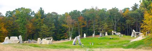 A panoramic view of large stone formations in a grassy area, surrounded by colorful autumn trees and people exploring.