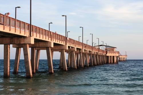 A wooden pier extends over calm blue waters, with a small building at the end and lights lining the walkway.