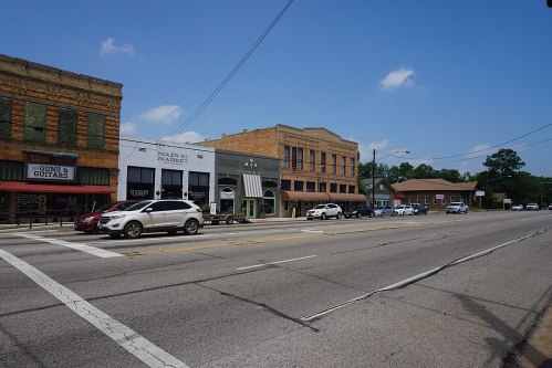 A small town street with shops, parked cars, and a clear blue sky. Historic buildings line the road.