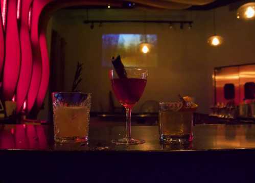 Three stylish cocktails on a bar, illuminated by soft red lighting, with a blurred background of a projected image.