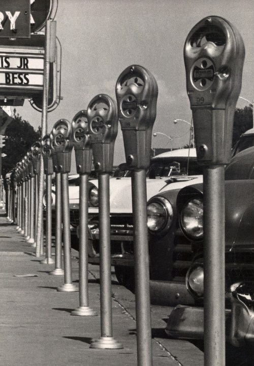 A row of vintage parking meters lined up along a sidewalk, with classic cars parked nearby.