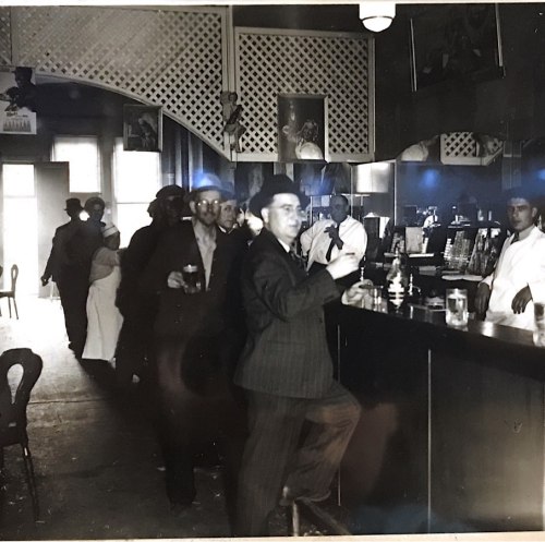 A black-and-white photo of men at a bar, some holding drinks, with a bartender serving in a vintage setting.
