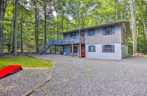 A two-story house surrounded by trees, featuring a gravel yard and a blue staircase leading to a deck.