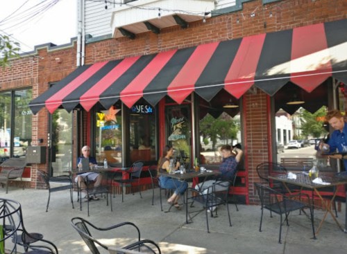 Outdoor seating at a restaurant with red and black awning; patrons enjoying drinks at tables on a sunny day.