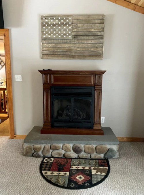 A rustic fireplace with a wooden mantel, stone base, and a wooden American flag art piece above it.
