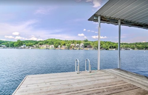 A serene lakeside view from a wooden dock, with houses nestled among green trees across the water.