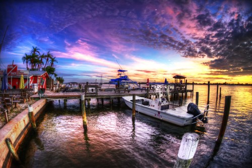A serene waterfront scene at sunset, featuring a docked boat, colorful sky, and palm trees along the shore.