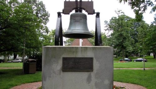 A large bell mounted on a stone pedestal in a park, surrounded by trees and grass.