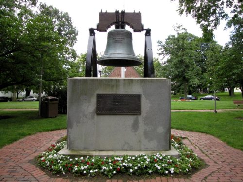 A large bell monument surrounded by flowers in a park, with trees and benches in the background.
