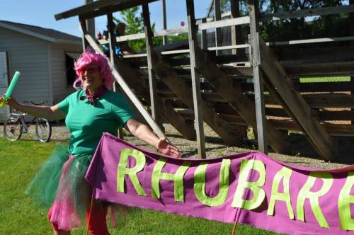 A person in a pink wig and colorful outfit holds a banner that says "RHUBARB" in a grassy area near bleachers.
