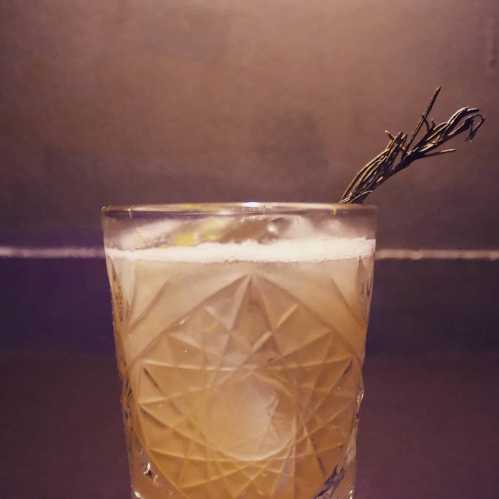 A close-up of a cocktail in a textured glass, garnished with a sprig of rosemary against a dark background.