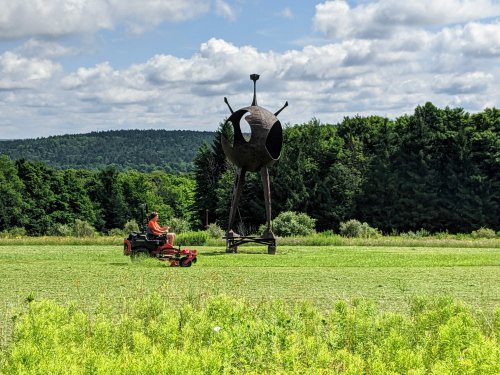 A person mows grass near a large, abstract metal sculpture in a green field with trees and clouds in the background.