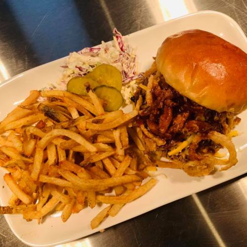 A plate featuring a pulled pork sandwich with onion rings, coleslaw, pickles, and a side of crispy French fries.