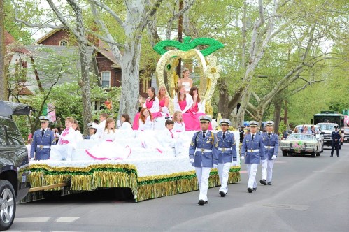 A parade float with young women in pink dresses, surrounded by greenery, while military personnel march alongside.