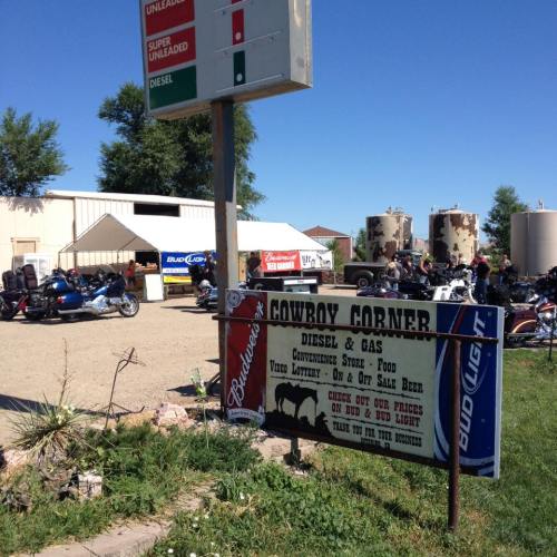 A gas station with a sign for "Cowboy Corner," surrounded by motorcycles and a food store, under a clear blue sky.