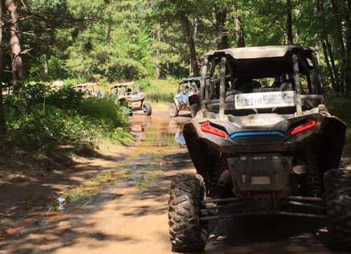 A line of off-road vehicles driving through a muddy trail in a wooded area.