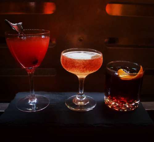 Three cocktails on a dark surface: a red drink in a tall glass, a light-colored drink in a coupe, and a dark drink with an orange twist.