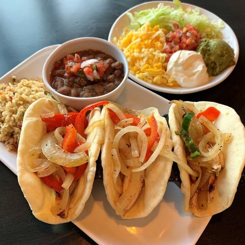 Three tacos filled with grilled chicken, peppers, and onions, served with rice, beans, and a side of toppings.