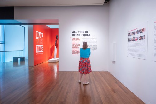 A visitor reads an exhibit titled "All Things Being Equal" in a modern art gallery with colorful walls.