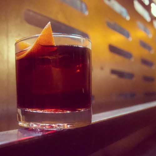 A glass of dark cocktail garnished with an orange twist, set against a warm, textured background.