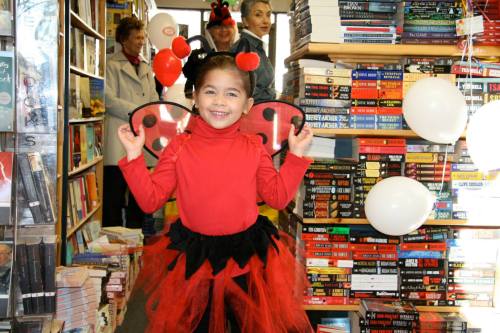 A smiling girl in a ladybug costume stands in a bookstore, surrounded by books and balloons.