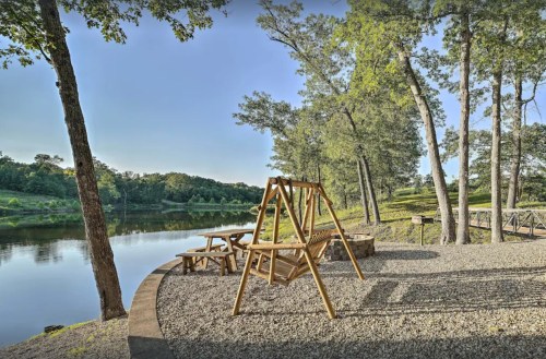 A wooden swing set and picnic table by a serene river, surrounded by trees and a clear blue sky.