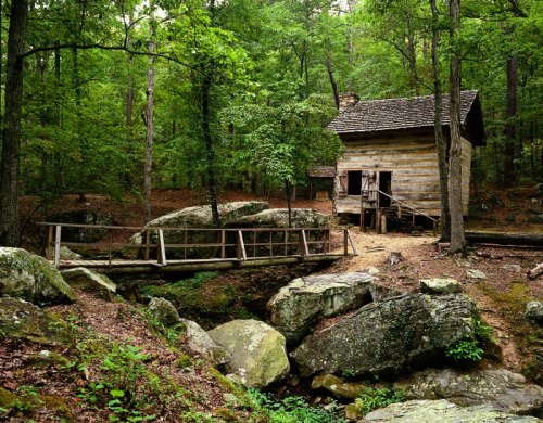 A rustic wooden cabin surrounded by lush green trees and large rocks, with a small bridge over a stream nearby.
