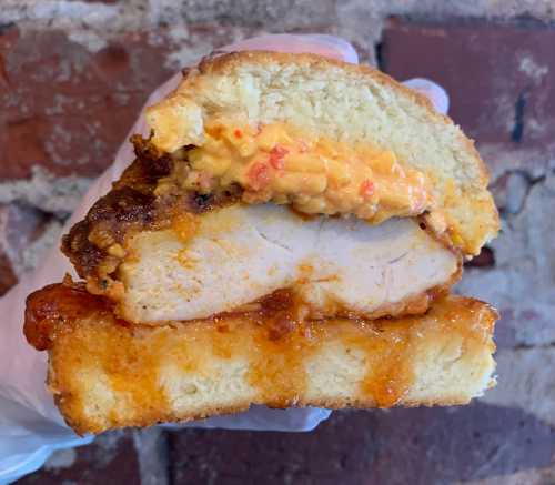 A hand holding a cross-section of a sandwich with fried chicken, pimento cheese, and a sweet sauce on a soft bun.