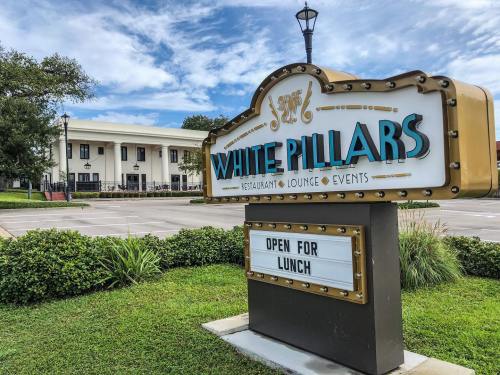 Sign for White Pillars restaurant and lounge, with "Open for Lunch" displayed, set against a blue sky and greenery.