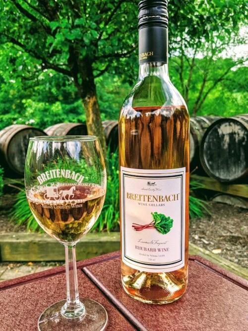 A bottle of Breitenbach Rhubarb Wine next to a glass filled with wine, set against a backdrop of wine barrels and greenery.