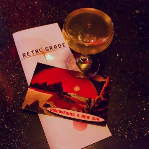 A cocktail sits on a bar next to a postcard titled "Discovering a New Sun" and a menu for Retrograde.