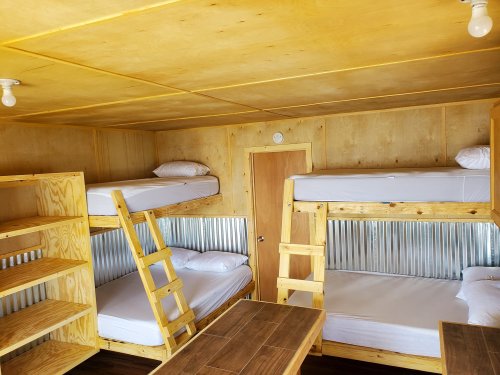 Interior of a cozy cabin with wooden bunk beds, a table, and simple decor, featuring warm yellow walls and metal accents.
