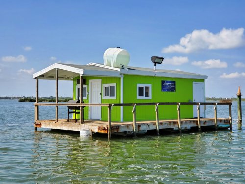 A bright green floating house on stilts in calm water, with a clear blue sky and a few clouds in the background.