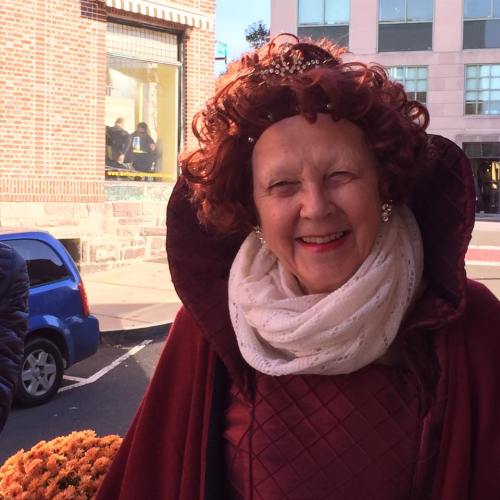 A smiling woman in a red costume and white scarf stands outdoors, with a city street and a car in the background.