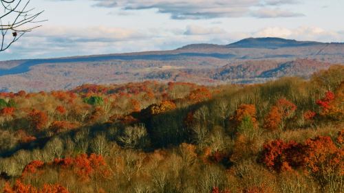 A scenic view of rolling hills covered in vibrant autumn foliage under a partly cloudy sky.