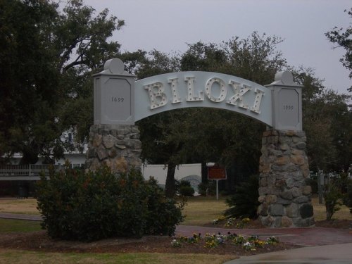 Sign archway reading "Biloxi" with stone pillars, surrounded by greenery and flowers, under a cloudy sky.