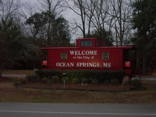 A red train caboose sign reading "Welcome to the City of Ocean Springs, MS," surrounded by trees and shrubs.