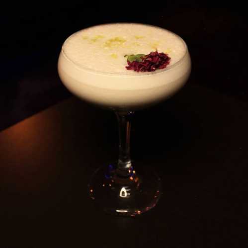 A frothy cocktail in a coupe glass, garnished with a flower and lime zest, set against a dark background.