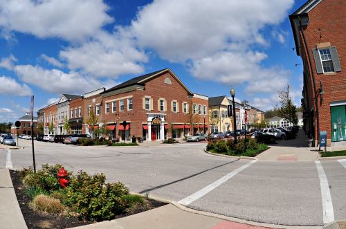 A vibrant town square with brick buildings, shops, and a clear blue sky dotted with clouds.