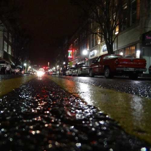 A wet street at night, reflecting lights from buildings and vehicles, with parked cars lining the sides.