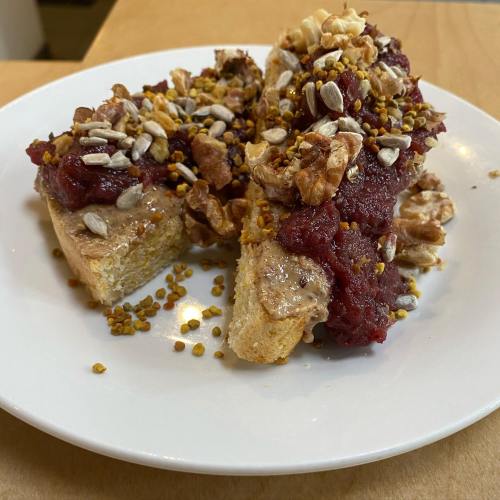 Two slices of bread topped with nut butter, red fruit spread, granola, sunflower seeds, and bee pollen on a white plate.
