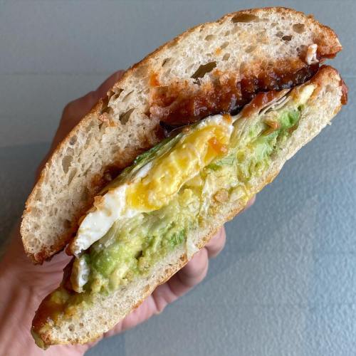 A hand holding a sandwich with layers of avocado, fried egg, and sauce between two slices of bread.