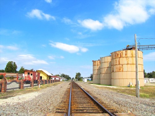 View of railroad tracks leading towards silos and buildings under a blue sky with scattered clouds.