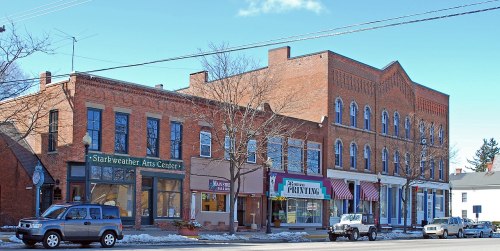 A street view of historic brick buildings, including an arts center and a printing shop, with parked cars and clear skies.