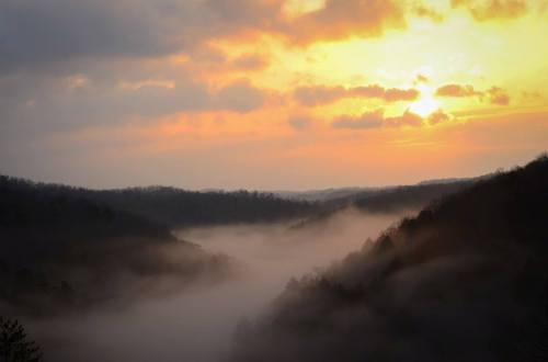 A serene landscape at sunset, with mist rolling through valleys and silhouetted hills under a colorful sky.
