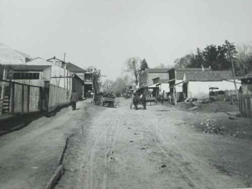 Black and white photo of a dirt road lined with buildings and horse-drawn carts in a rural setting.
