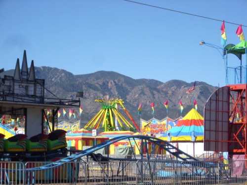 Colorful carnival rides and tents set against a backdrop of mountains under a clear blue sky.