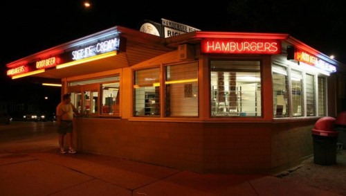 A brightly lit hamburger stand at night, featuring neon signs and a customer ordering at the window.