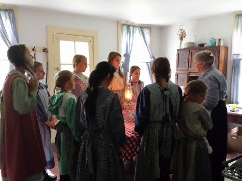 A group of girls in period dresses gathers around a table with a lantern, listening to an older woman in a historical setting.