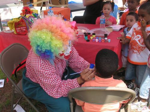 A clown with a colorful wig paints a child's face at a festive outdoor event, surrounded by curious children.
